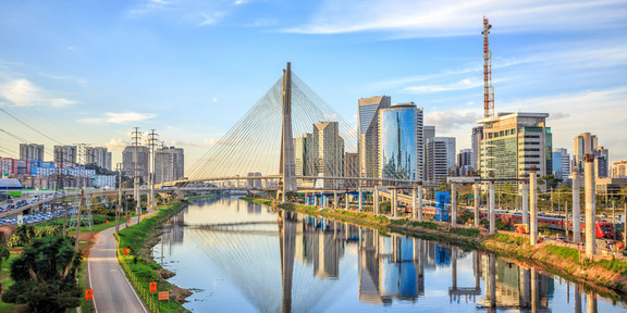 Photo: The “Octavio Frias de Oliveira” bridge in São Paulo spans a shimmering river. Next to the bridge are green parks and gleaming high-rise buildings with glass fronts.