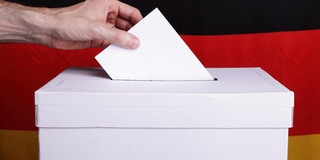Eine Person wirft einen Wahlzettel in eine Wahlurne. Im Hintergrund ist eine Deutschlandfahne