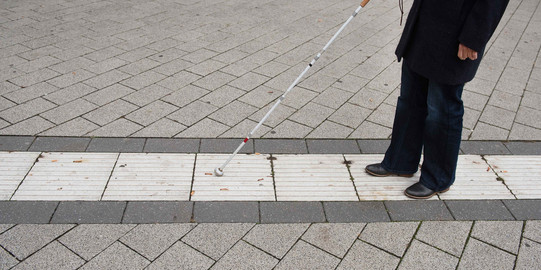 Blindenleitsystem Eine blinde Person folgt dem Blindenleitsystem auf dem Campus mithilfe eines Blindenstocks